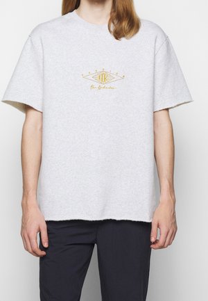 Camiseta de algodón gris con un ajuste relajado, mangas cortas y un dobladillo sin rematar. Presenta un logo bordado en oro en la parte delantera.