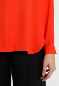 Blouse légère rouge à manches longues, ourlet arrondi et poignets boutonnés, associée à un pantalon noir. Texture lisse, détails minimalistes.