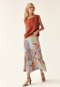 Haut sans manches en lin couleur rouille associé à une jupe maxi bleu clair ornée de motifs de feuilles de palmier rouges et jaunes. Sandales à talons marron.
