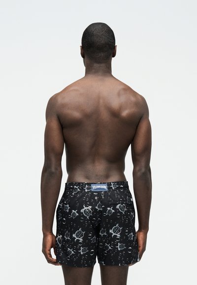 Άνδρας που στέκεται γυρισμένος προς τα πίσω, φοράει μαύρο μαγιό με σχέδια λευκής θαλάσσιας χελώνας και φυσαλίδες, σε απλό ανοιχτό φόντο.