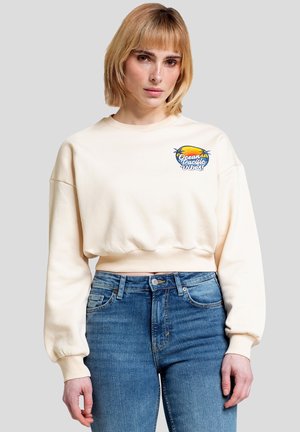 Kvinde med blond bob-frisure iført en cremefarvet cropped sweatshirt med "Ocean Pacific Vibes" logo og blå højttaljede jeans, stående mod grå baggrund.