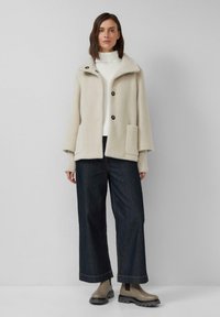 Beige, gestructureerde wollen jas met een kraag, knoopsluiting en een voorzak, gecombineerd met donkere wijd-uitlopende jeans en enkellaarsjes.