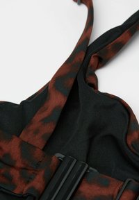 Tkanina s černými a rezavými vzory s texturovaným designem. Nastavitelné popruhy a detail v podobě obdélníkového černého kovu. Měkký a flexibilní materiál.