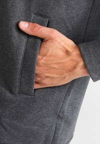 Prenda texturizada de color gris con un bolsillo lateral. Una mano está colocada dentro del bolsillo, mostrando un diseño ajustado.