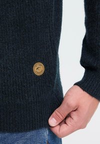 Kootud tume roheline sviiter ribade struktuuriga, millel on ümar pruun logo märk vasakul all nurgas ja lõdvem lõige.