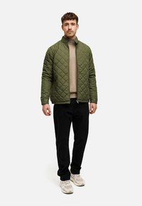 Quiltet olivengrøn jakke med ståkrave, en lynlås foran, båret over en beige turtleneck, parret med sorte bukser og lyse sneakers.