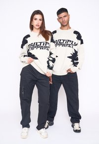 Două modele poartă pulovere crem asortate cu modele abstracte negre și textul "MULTIPLY APPAREL", combinate cu pantaloni cargo și blugi negri.