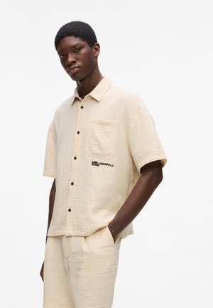 Junger Mann trägt ein beiges, kurzärmeliges Hemd zum Knöpfen und passende Shorts mit dem "Karl Lagerfeld Jeans" Logo auf der Hemdtasche.