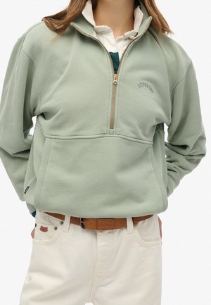 Osoba ubrana w jasnooliwkową bluzę z suwakiem do wysokości klatki piersiowej, na białą koszulę i kremowe spodnie z brązowym skórzanym paskiem.