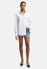 Chemise blanche à boutons avec un col, des manches longues et une coupe décontractée, associée à un short en denim et des escarpins noirs à bout pointu.