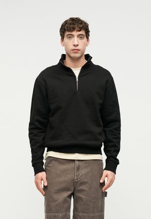 Młody mężczyzna zwrócony przodem, ubrany w czarny pulower na suwak o długości ćwiartki, na kremową koszulę i szare spodnie, na tle jednolitego jasnego tła.