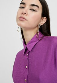 Camisa color púrpura con collar y un acabado texturizado, que presenta un cierre frontal de botones. Pendientes metálicos dorados con una forma orgánica. Fondo minimalista.