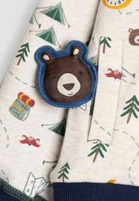 Krēmpalvas audums ar kempinga motīviem, ieskaitot teltis, kokus un brūna lāča pašu. Zils kontūrs ap lāci izceļ tā seju.