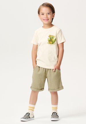 Creamfarbenes T-Shirt mit Obstgrafik, kombiniert mit olivgrünen strukturierten Shorts. Schwarz-weiße karierte Slipper mit gelb gestreiften Socken.