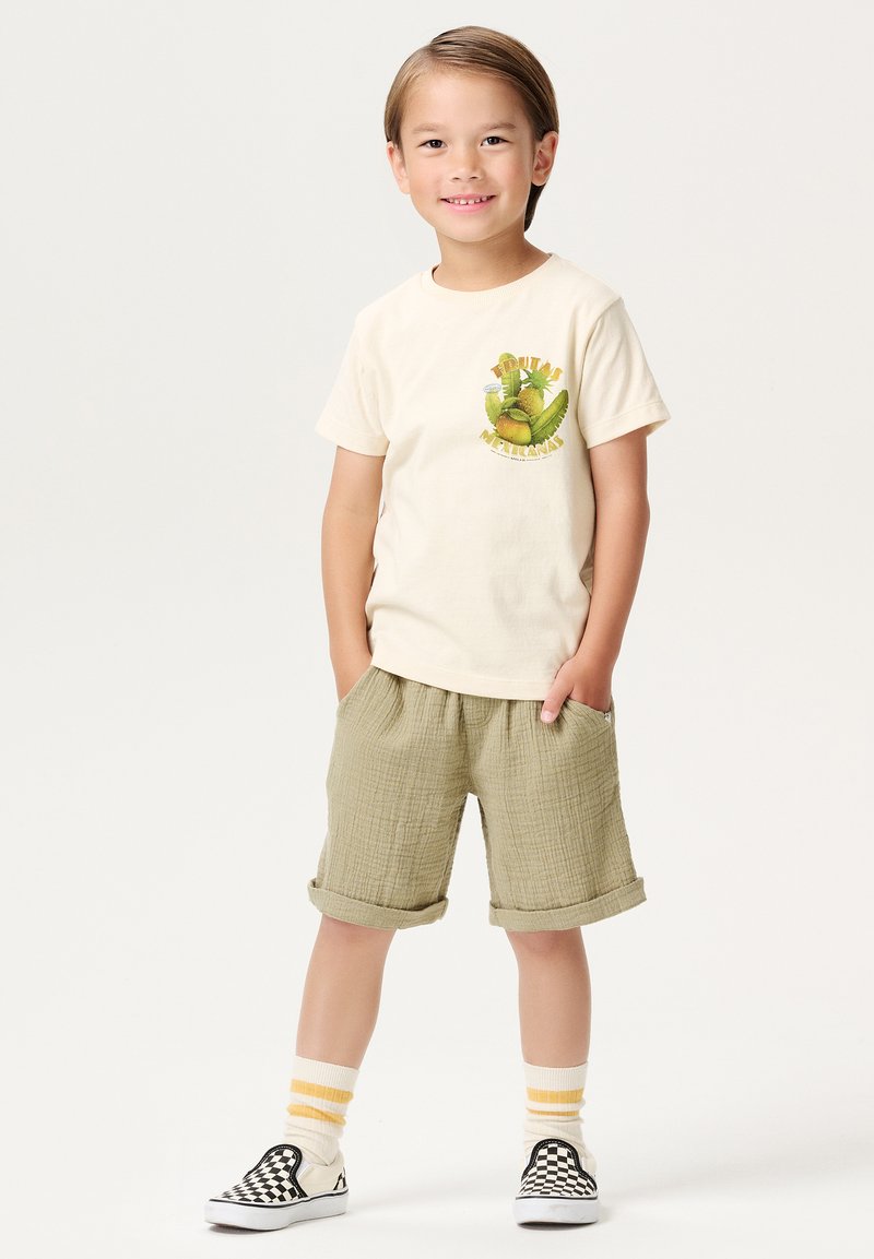 Creamfarbenes T-Shirt mit Obstgrafik, kombiniert mit olivgrünen strukturierten Shorts. Schwarz-weiße karierte Slipper mit gelb gestreiften Socken.