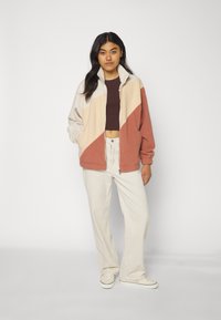 Chaqueta de forro polar con cremallera en beige, crema y óxido, con cuello alto, bolsillos laterales y puños acanalados, combinada con pantalones de pana blancos.