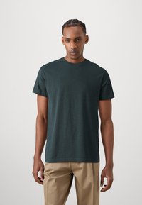 Dunkelgrünes T-Shirt mit Rundhalsausschnitt und kurzen Ärmeln, aus weichem Material; locker sitzend mit geradem Saum; kombiniert mit hellbeigen Hosen.