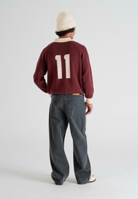 Man wearing a maroon sweater with number 11 on the back, dark wide-leg jeans, white beanie, and white sneakers standing facing away.