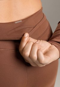 Brązowe, rozciągliwe legginsy sportowe z widocznym logo na pasku, o dopasowanym kroju i gładkiej teksturze. Ręka trzymająca materiał.