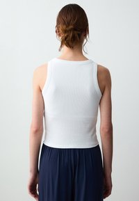 Donna con capelli castani raccolti in uno chignon, che indossa un top bianco a costine senza maniche e pantaloni larghi blu scuro, in piedi di spalle su uno sfondo neutro.