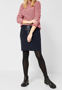 Růžový svetr s texturou a tříčtvrtečními rukávy spojený s tmavě modrou sukní se stahovací šňůrkou v pase a dvěma bočními kapsami. Černé kotníkové boty.