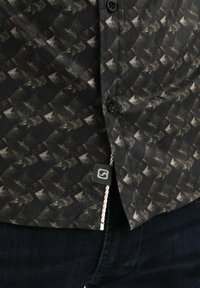 Camisa de botones de manga corta con tejido negro y un patrón geométrico en tonos de marrón y verde; incluye una etiqueta de la marca en el dobladillo.