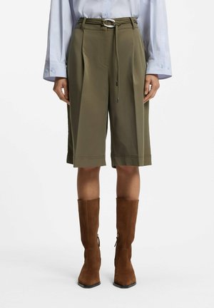 Persoon draagt olijfgroene wijde knielange shorts met riem, wit hemd met lange mouwen en bruine kniehoge suède laarzen op witte achtergrond.