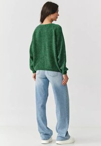 Zielony, puszysty sweter z szerokim dekoltem i luźnymi rękawami, w połączeniu z jasnoniebieskimi dżinsami o prostym kroju i białymi sneakersami.