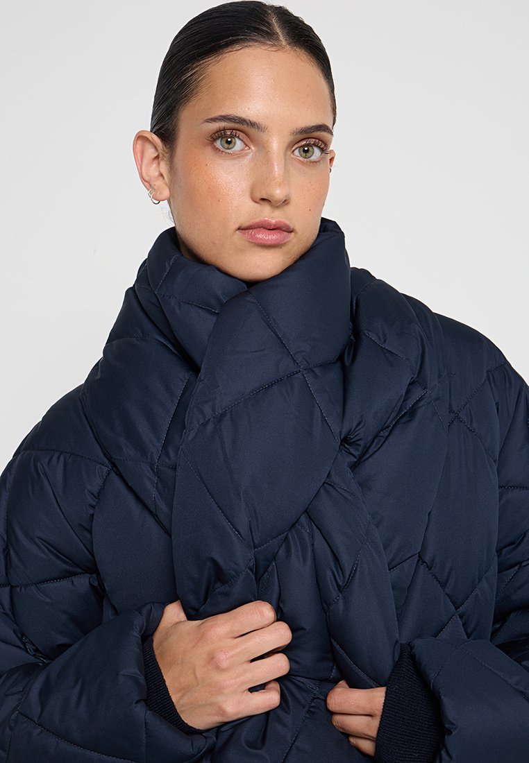 Granatowa kurtka puchowa o pikowanej fakturze, z założonym kołnierzem i prążkowanymi mankietami. Posiada wysoki krój kołnierza dla dodatkowego ciepła i osłony.