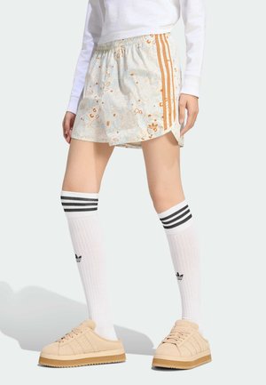 Person trägt Shorts mit Blumenmuster und orangen Streifen, lange weiße Socken mit schwarzen Streifen und beige Turnschuhe vor hellem Hintergrund.