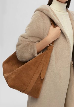 Donna che indossa un cappotto di pelliccia beige e un maglione a collo alto bianco, mentre porta una grande borsa a tracolla in suede marrone con tracolla regolabile.
