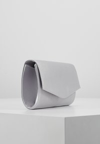 Pochette en satin argenté avec un rabat triangulaire, debout sur une surface blanche contre un fond gris uni.