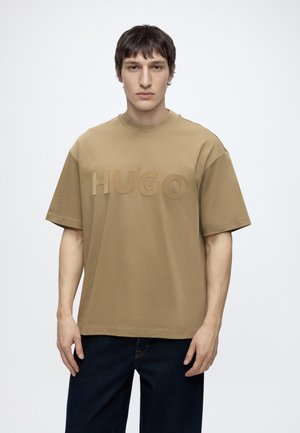 Young man with short dark hair wears loose beige T-shirt with embossed "HUGO" logo and dark jeans, standing against plain light background.