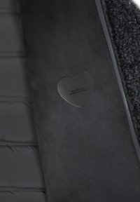 Giacca nera con texture che presenta una sezione imbottita, tessuto liscio e un'etichetta a forma di cuore con il testo "Realizzato con amore, progettato in Toscana."