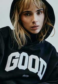 Schwarzer Hoodie mit großer weißer Blockschrift "GOOD" auf der Vorderseite. Weicher Stoff, lässige Passform, mit Kapuze und dezente Texturdetails.