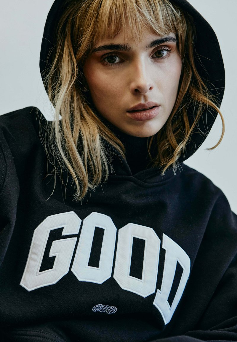 Schwarzer Hoodie mit großer weißer Blockschrift "GOOD" auf der Vorderseite. Weicher Stoff, lässige Passform, mit Kapuze und dezente Texturdetails.
