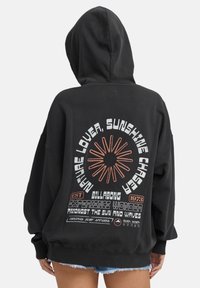 Czarna bluza z kapturem z dużą grafiką na plecach, z pomarańczowym i białym tekstem, wzór promieni słonecznych oraz luźny krój z kieszenią z przodu.