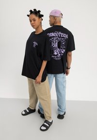 Twee modellen dragen zwarte oversized "Discoteca" grafische t-shirts, één kijkt naar voren met beige broek en sandalen, de ander kijkt naar achteren met lichte spijkerbroek en een pet.