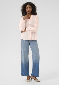 Chemise boutonnée rose clair en tissu texturé à motifs ; associée à un jean bleu à larges jambes et des escarpins beiges.