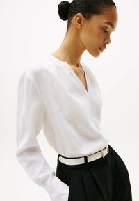 Blusa de lino blanca con escote en V, mangas largas y pliegues sutiles en los hombros, combinada con pantalones negros de talle alto y un cinturón blanco.