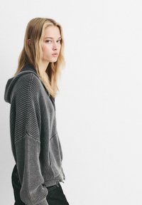 Grå strikket hættejakke med et tekstureret mønster og snoretræk i hætten. Har ribbede manchetter og en let slidt kant. Bæres med mørke jeans.