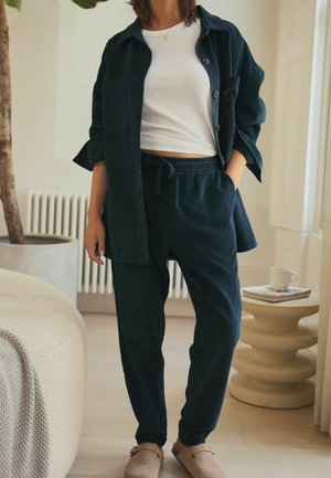 Woman wearing dark blue corduroy button-up shirt and matching pants with white cropped top and beige slip-on shoes, standing indoors near table.