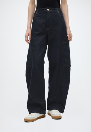 Person trägt schwarze Cargo-Jeans mit weitem Bein und Seitentaschen sowie weiße Sneaker, steht vor einem einfarbigen hellen Hintergrund.