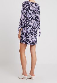 Kobieta stoi, widoczna od tyłu, ubrana w sukienkę w kwiatowy wzór z długimi rękawami i zawiązywaniami przy mankietach, do której dobrano białe szpilki.