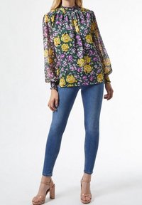 Blusa floreale con fiori gialli e viola su sfondo verde, maniche lunghe trasparenti con polsini arricciati, abbinata a jeans skinny blu e décolleté beige.