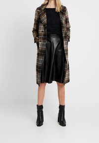 Rutig trenchcoat i bruna och krämfärgade nyanser över en svart texturerad tröja, kombinerad med en svart läderskjorta och svarta klackiga stövlar.