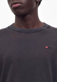 Jeune homme portant un t-shirt gris foncé Diesel à col rond avec un petit logo rouge et blanc sur la poitrine gauche.