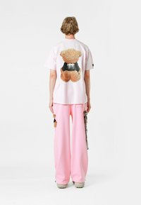 Camiseta de manga corta rosa con un gráfico de un oso de peluche en la parte posterior. El modelo la combina con pantalones rosa de pierna ancha y calzado texturizado.