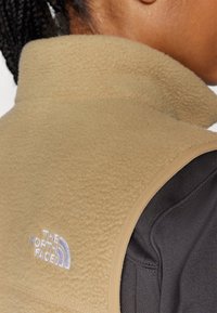 Beige fleece vest met hoge kraag en zwarte accenten, met het "The North Face" logo in witte borduurlogo op de achterkant. Textuurdetail van de stof zichtbaar.