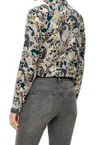 Blusa con motivo floreale color crema, con accenti in teal, marrone e giallo. Maniche lunghe, collo a camicia, vestibilità leggermente rilassata. Abbinata a jeans grigi.